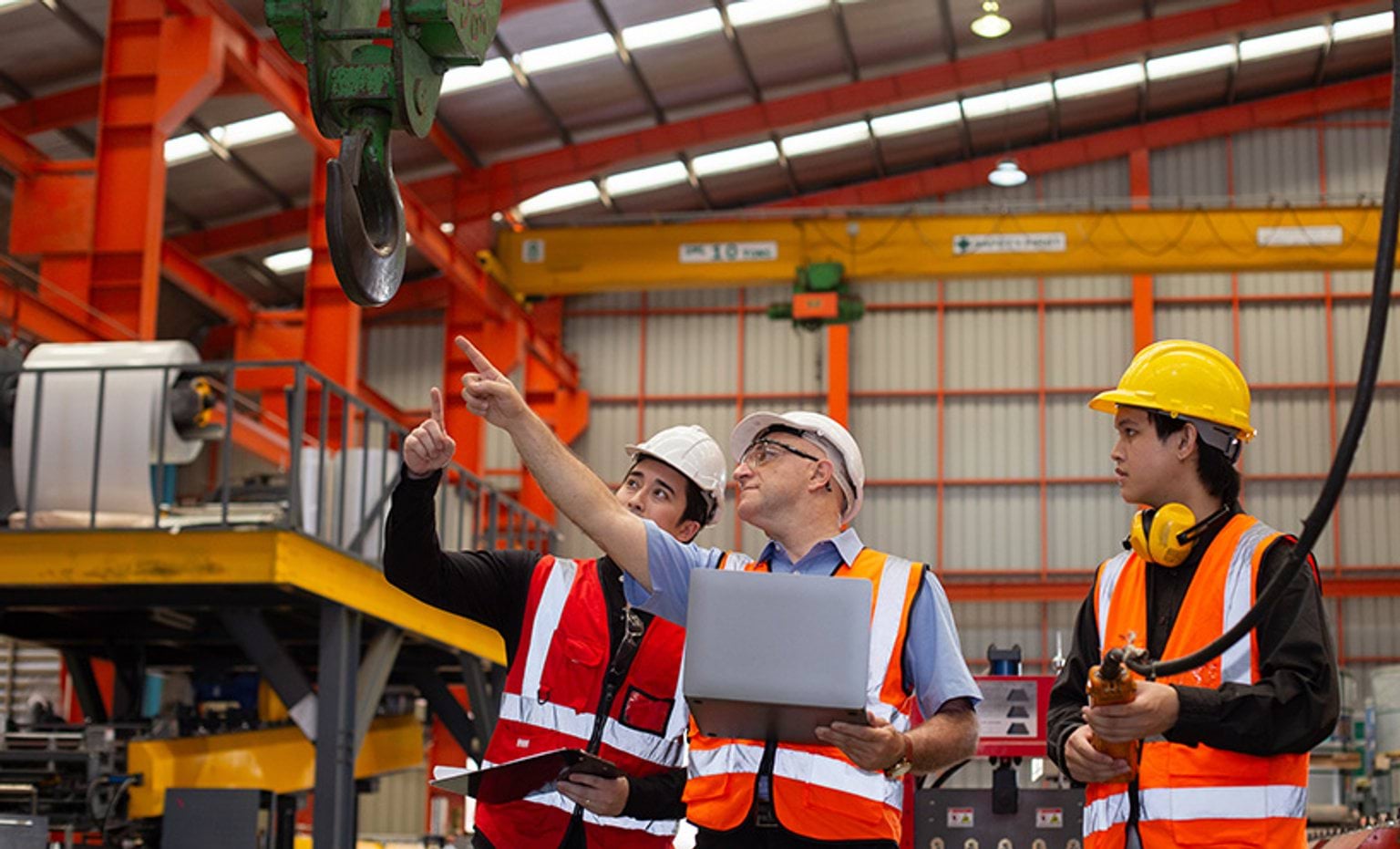 factory workers wearing safety gear analyse overhead crane in manufacturing plant for workplace safety.